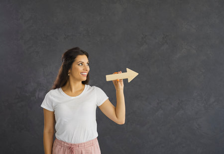 Young caucasian woman standing on a gray background holding a wooden arrow pointing to the side. Smiling brunette woman in casual clothes looking at free space for text. Concept of the right directionの写真素材