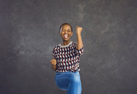 Joyful emotional afro american woman dancing and having fun celebrating victory and success. Cheerful woman laughs out loud, clenched her fists happily and jumps on a gray background.の写真素材