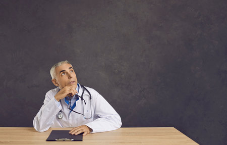 Portrait of dreamy or thinking senior male doctor with pensive expression on gray banner background. Man in medical uniform sits at table and, holding his chin, looks intently at free space for text.の写真素材