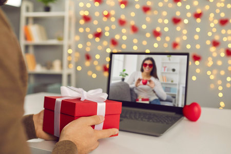 Close up of red gift box in male hands in front of laptop screen during online date. Man shows the gift he prepared for Valentines Day. Concept of distance relations. Selective focus.の写真素材