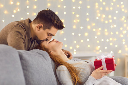 Side view of a loving husband kissing his young wife on the forehead after giving her a gift. Man tenderly greeted the woman with her special date. Couple in love celebrating at home in a cozy room.の写真素材
