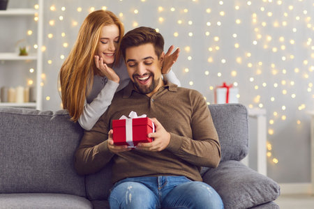 Joyful excited man sits on the sofa in the room and receives a gift from his girlfriend. Young woman congratulated her boyfriend on Valentines Day, Christmas, or his birthday. Holiday concept.の写真素材