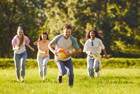 Happy joyful young people friends in casual clothes playing with a ball on a green grass in the park outdoors. Men and women running on the lawn with ball and having fun in summer.の写真素材