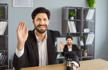 Cheerful man dressed in formal suit, shooting video blog on camera while posig at workplace, waving hello to followers, having online chat, smiling happily, posing against office interiorの写真素材