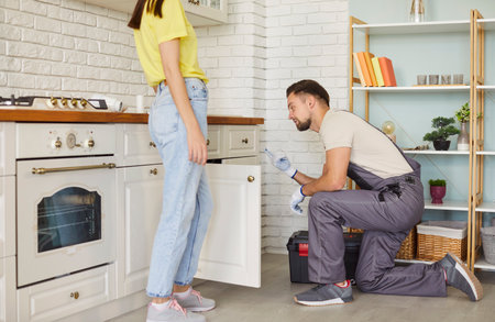 Professional man plumber fixing problems with sink, clogged drain, woman client standing near, explaining what is wrong, supplying good quality. Maintenance, housekeeping and household service.の写真素材
