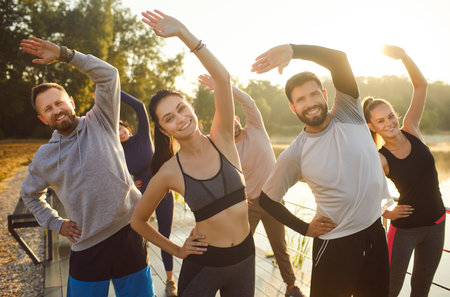 Group of four young people participating in fitness class outdoor, raising their arms, doing exercise routine, such as yoga, aerobics, or stretching session in nature. Sport, healthy lifestyle conceptの写真素材