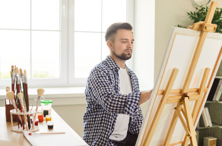 Young 25s European male artist painting on easel at home studio or workshop, looks focused on work or hobby, bringing his creative vision to life. Talent, inspiration and passion conceptの写真素材