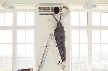 Service worker wearing overalls, standing on ladder, repairing air conditioner, checking if there are any problems, posing in spacious room. Maintenance and repair service concept.の写真素材