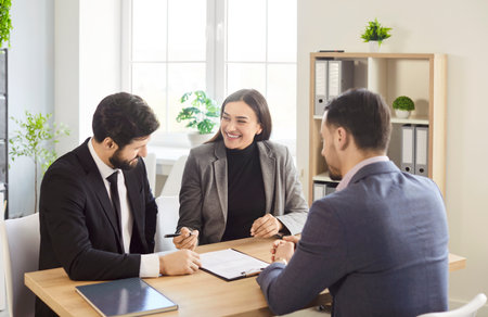 Portrait of cheerful happy young couple sitting in office with male agent going to sign a contract. Man and woman reaching agreement with realtor or financial advisor sitting at desk on workplace.の写真素材
