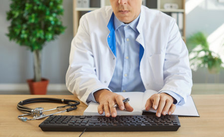 Closeup of professional male medical doctor in white uniform working at the desk in office typing on pc computer on his workplace. Physician man working in clinic. Medicine concept.の写真素材