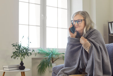 Serious senior woman covered in cozy blanket sitting on home sofa and calling on mobile phone. Old person with glasses holding plaid, grandmother using smartphone to call. Talk, connection conceptの写真素材