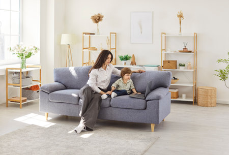Family of mother and son, babysitter, cute little kids children enjoy using laptop watching cartoons, internet video call or shopping online looking at computer screen sitting together on sofa at homeの写真素材