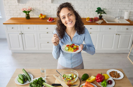 Portrait of a happy cheerful young woman holding plate with fresh healthy vegetable salad and looking at camera standing in kitchen at home. Vegetarian food, diet and nutrition concept.の写真素材