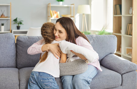 Portrait of happy smiling woman housewife hugging her cute little child daughter holding freshly washed towel in hands sitting on sofa in living room at home. Laundry and family housework concept.の写真素材