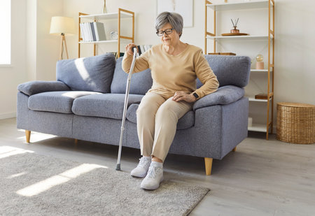Senior old woman at home alone, mature female with grey hair on sofa leaning on stick feeling hard to move, sad tired in need of rehabilitation after leg trauma, grandmother having walking disabilityの写真素材