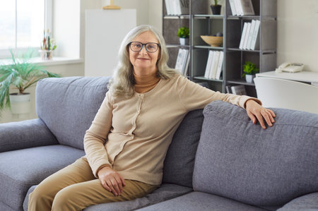 Happy senior woman sitting on sofa to enjoy cozy time in home living room. Calm cheerful elderly lady in glasses relaxing with smile and positive optimistic look. Wellbeing, retirement conceptの写真素材