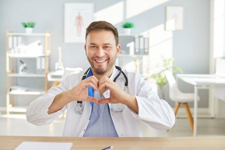 Smiling male doctor cardiologist in white coat sitting at desk in medical office, making heart shape with his hands. Cheerful healthcare professional expressing love, care and support during workday.の写真素材