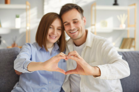 Couple in love making finger heart frame, loving portrait, happy man and woman in love, having fun, posing, smiling, enjoying being together, celebrating holiday together, sitting close at sofaの写真素材