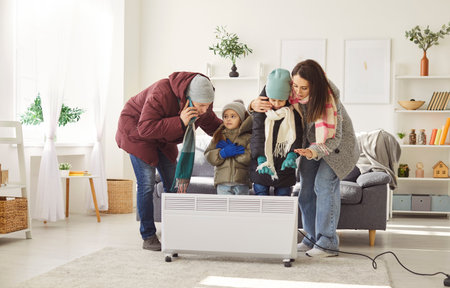 Cold parents and children dressed in warm clothes standing by electric heater, trying to get warm. Family experiencing heating issues at home, feeling chill and calling emergency services for help.の写真素材