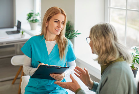 Smiling nurse in blue uniform with clipboard listening to senior woman during medical consultation near bright window. Friendly healthcare professional engaging in warm conversation with patient.の写真素材
