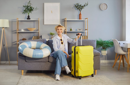 Portrait of a happy woman with suitcase sits on sofa at home living room, ready for summer travel trip in a cozy setting with focus on vacation preparation and excitement before departure.の写真素材