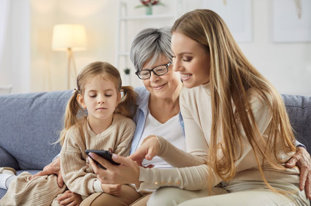 Daughter and granddaughter teaching senior mother or grandmother how to use smartphone, using wireless tech, download app for chatting, choosing what to watch online. Family and pastimeの写真素材