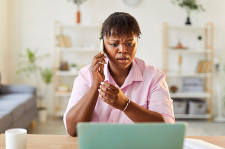 African American woman who has suffered from online fraud and Internet scam is looking at laptop, having problem with bank account, holding mobile phone, speaking to customer service helpline managerの写真素材