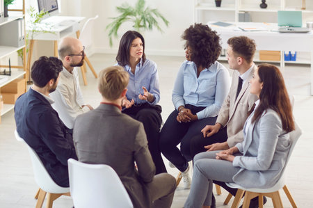 Group of multiracial company employees sitting together in circle during business meeting or training, listening to their colleague or business trainer. Corporate culture and team building concept.の写真素材