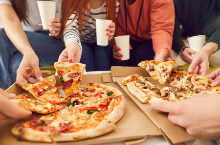 Close up hands of a young people friends gathering in pizzeria or at home together eating tasty Italian food taking pizza slices from box enjoying party with cardboard cups in hands.の写真素材