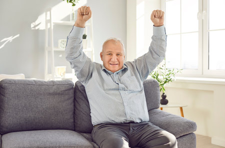 Elderly excited senior man sitting on sofa in living room at home holding hands up in fist like a winner. Happy smiling retired person wearing blue shirt looking at camera with satisfied expression.の写真素材
