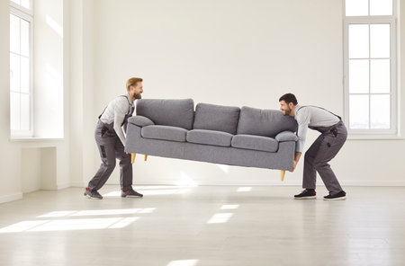 Two movers in uniform lifting large sofa and bringing it into bright empty room. Professional workers assisting with furniture transportation during home relocation or delivery service for client.の写真素材