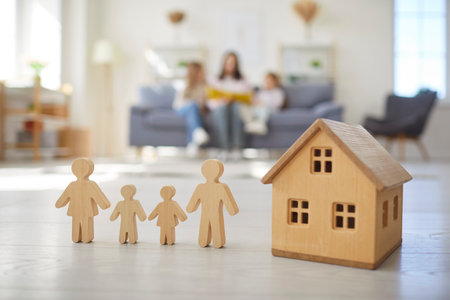 Little wooden toy house on floor with figures of people in living room at home with family of mother and children sitting together on sofa in background. Real estate and insurance conceptの写真素材
