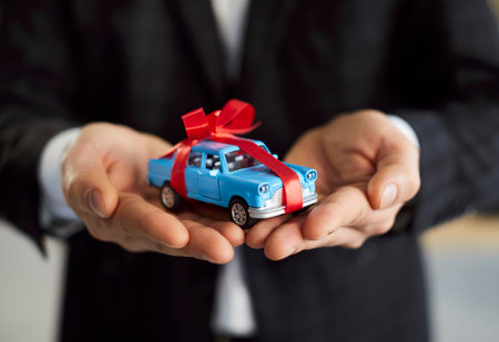 Cropped closeup photo of man seller in formal black suit holding small toy car with red gift bow in...