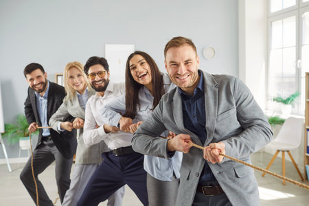 Cheerful multicultural business team playing tug of war together in office. Smiling colleagues pulling rope during teamwork activity, showing unity, energy, motivation and collaboration spirit.の写真素材