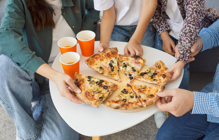 Cropped top view of family members reaching for slices of hot pizza on wooden plate over white table with orange paper cups nearby. Parents and children enjoying delicious food together during dinner.の写真素材