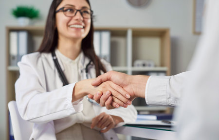 Smiling female doctor in white coat shaking hands with patient in medical office. Professional friendly physician greeting or congratulating client, showing support and care. Cropped shot. Low angle.の写真素材