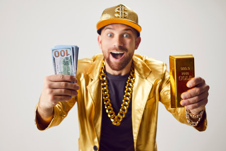 Surprised, rich man in a golden suit holding a stack of cash money and a gold bar against a white...