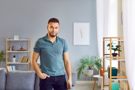 Young confident attractive dark-haired bearded man posing in casual clothes holding hand in pocket of trousers. Portrait of handsome man wearing shirt and jeans, looking at camera in the living room.の写真素材