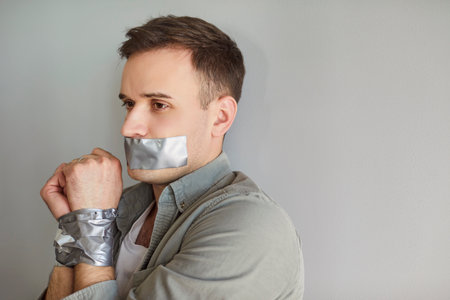 Young man with mouth and hands taped, maintaining silence. Person appears distressed and constrained, possibly indicating a situation of restriction or secrecy.の写真素材