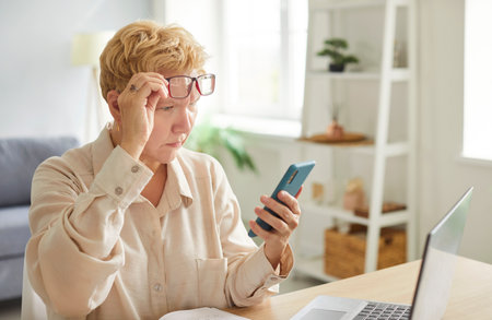 Senior woman pensioner sitting at the desk working with laptop at home and looking at mobile phone...