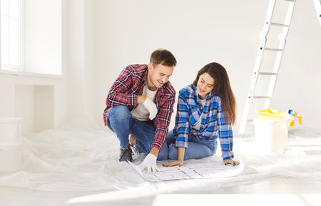 Happy young couple sitting on floor with blueprint in new apartment and discussing design project of...
