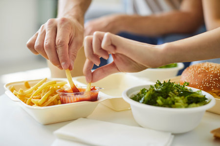 Unrecognizable couple eating together fast food, close-up cropped view, two hands dipping French...