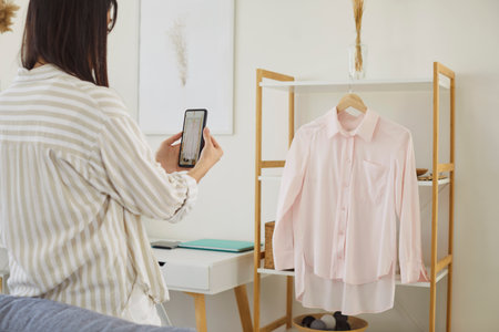 Young woman holding her smartphone to take photo of casual pink blouse hanging on hanger, preparing to sell clothes through online marketplace or promote it on social media as influencerの写真素材