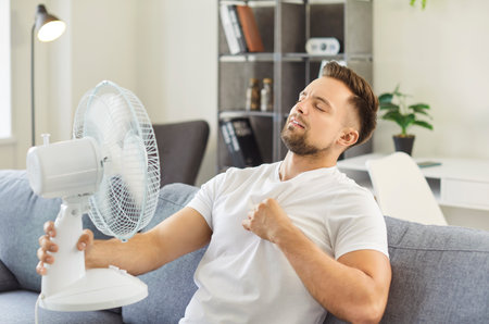 Man sitting on sofa feels discomfort from heat, using fan device blowing cool air, eyes closed, seeking relief in hot living room coping with high temperature, summer problem in overheated roomの写真素材