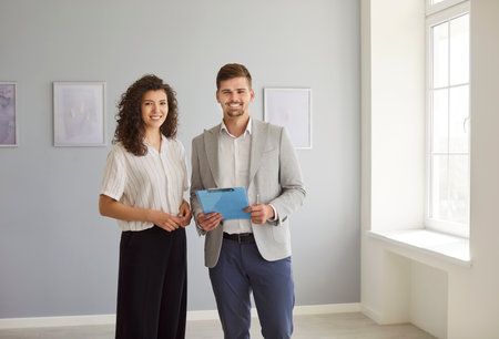 Smiling real estate agent standing with happy female in empty bright apartment. Portrait of cheerful...