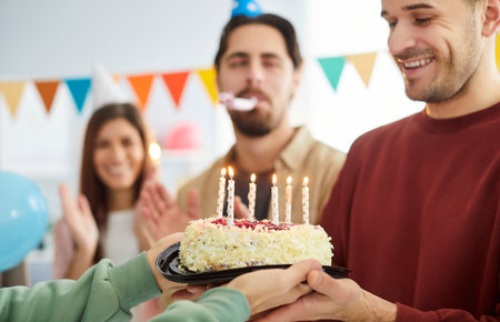 Close up of a smiling young happy man holding his cake with candles standing in the living room with...