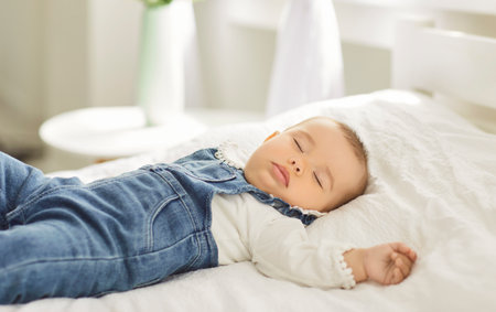 Peaceful adorable cute infant baby sleeping on white comfortable bed during daytime sleep. Portrait of little calm toddler child in denim overalls napping alone, resting in bedroom. Babyhood concept.の写真素材