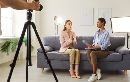 Businesswoman giving interview to journalist in front of camera sitting on sofa and giving talk in...