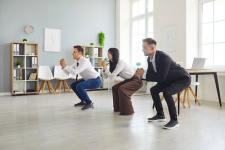 Three colleagues in business attire perform synchronized squats in a bright modern office, taking an...