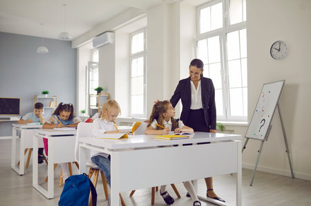Female friendly smiling teacher is helping cute little schoolgirl during lesson in elementary...